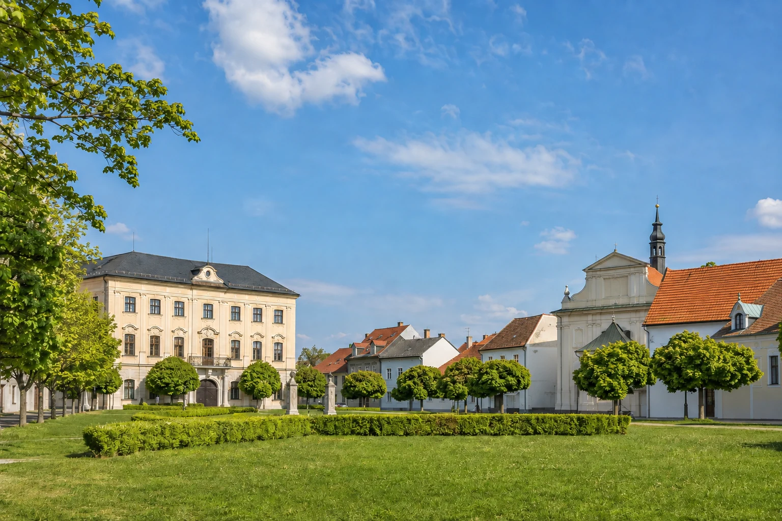 Historischer Ortskern von Hostivice mit hellem Schlossbau, Kirche, Wohnhäusern und begrüntem Platz